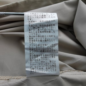 May include: A white rectangular garment care label with black Japanese text sewn onto a beige fabric surface. The label contains detailed washing instructions and manufacturing information, positioned against a smooth, light-colored textile background.