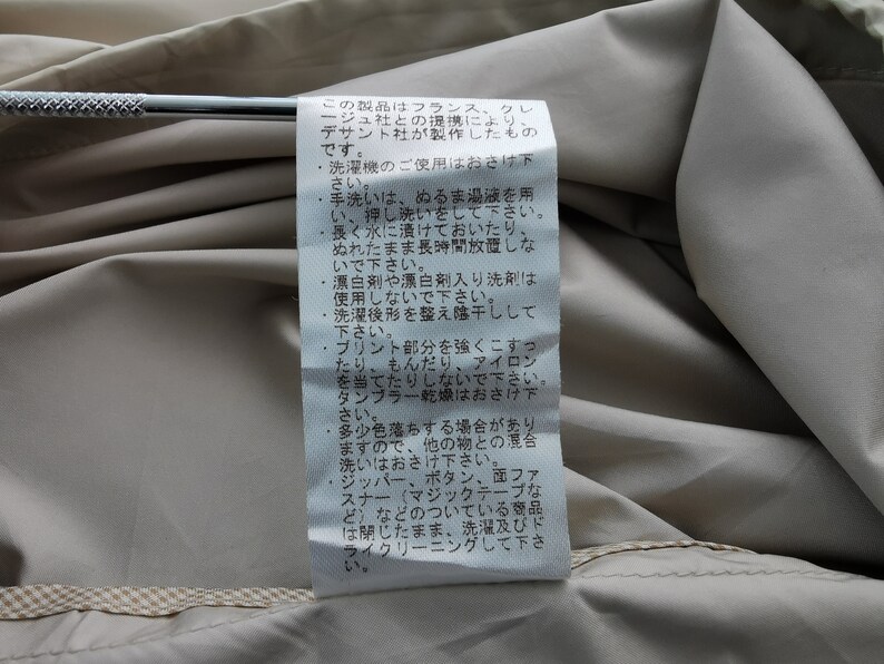 May include: A white rectangular garment care label with black Japanese text sewn onto a beige fabric surface. The label contains detailed washing instructions and manufacturing information, positioned against a smooth, light-colored textile background.