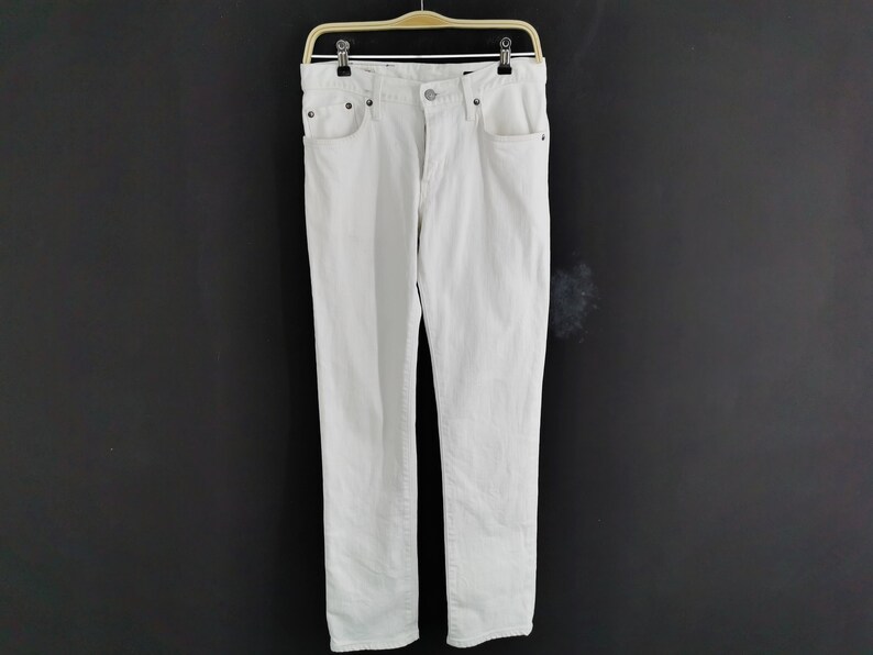 K&ouml;nnte beinhalten: Eine wei&szlig;e Jeans aus Denim, die an einem Holzb&uuml;gel vor einem dunklen Hintergrund h&auml;ngt. Die Jeans hat ein klassisches F&uuml;nf-Taschen-Design mit Knopf- und Rei&szlig;verschluss. Der Stoff scheint mittelschwer zu sein.