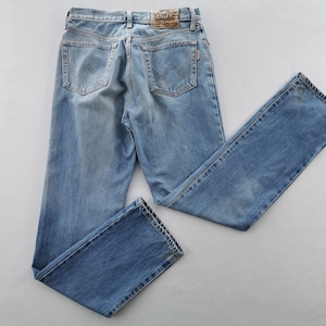Può includere: Jeans di denim blu chiaro con un classico design a cinque tasche. I jeans presentano un lavaggio sbiadito e una toppa in pelle con il marchio "EDWIN" sul retro. I jeans sono stesi su una superficie grigio chiaro.