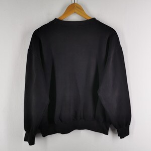 K&ouml;nnte beinhalten: Ein einfarbiges schwarzes Sweatshirt h&auml;ngt an einem Holzb&uuml;gel vor einer wei&szlig;en Wand. Das Sweatshirt hat einen Rundhalsausschnitt, lange &Auml;rmel und gerippte B&uuml;ndchen und Saum. Das Kleidungsst&uuml;ck scheint aus einem weichen, bequemen Material zu sein.