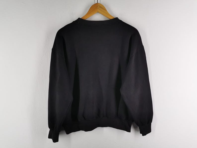 K&ouml;nnte beinhalten: Ein einfarbiges schwarzes Sweatshirt h&auml;ngt an einem Holzb&uuml;gel vor einer wei&szlig;en Wand. Das Sweatshirt hat einen Rundhalsausschnitt, lange &Auml;rmel und gerippte B&uuml;ndchen und Saum. Das Kleidungsst&uuml;ck scheint aus einem weichen, bequemen Material zu sein.