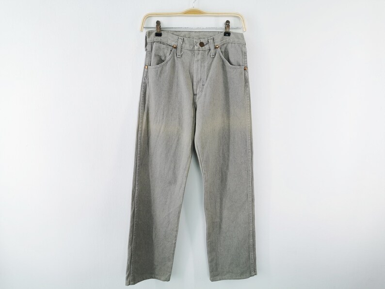 May include: Light gray denim jeans hanging on a gold-colored hanger. The jeans have a classic five-pocket design with a button and zipper closure. The pants are a straight-leg style.