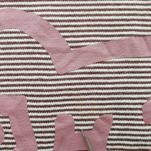 Pode incluir: Close-up de um tecido com um padr&atilde;o de riscas castanhas e brancas. Sobrepostos est&atilde;o v&aacute;rios peda&ccedil;os de tecido rosa cortados em formas abstratas. Uma tesoura &eacute; vis&iacute;vel.