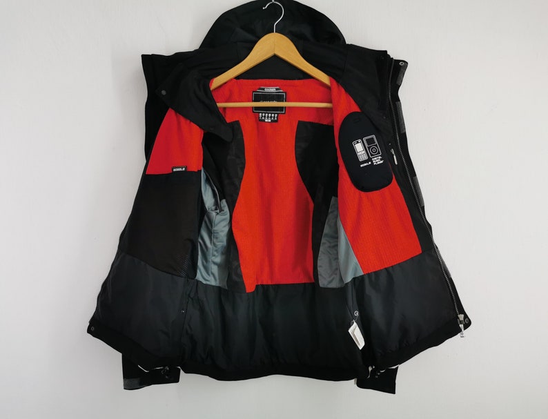 Pode incluir: Casaco de inverno preto com capuz, pendurado num cabide de madeira. O interior do casaco &eacute; vermelho brilhante, com bolsos cinzentos e pretos. O casaco est&aacute; aberto, revelando o forro interior e os bolsos. O casaco tem fecho de correr.