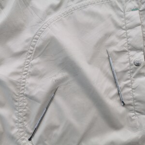 May include: A light beige nylon jacket featuring a curved seam detail, a vertical pocket with a silver-toned metal snap button, and two thin silver metal tools placed on the fabric surface to indicate a small area of wear or discoloration.