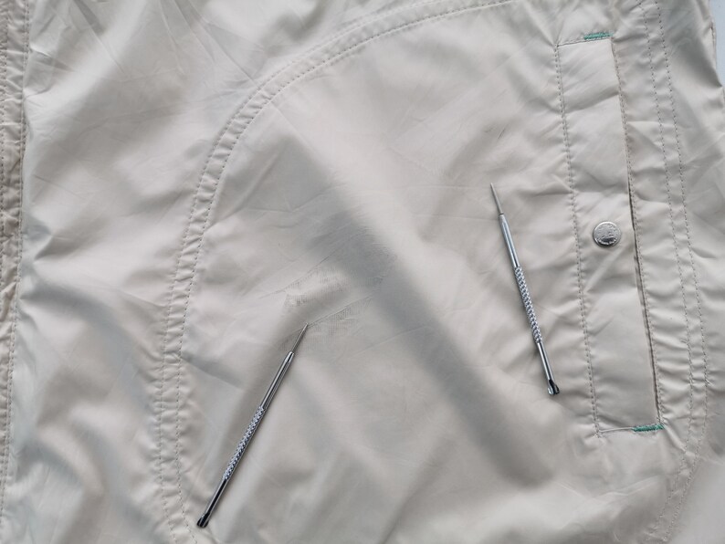 May include: A light beige nylon jacket featuring a curved seam detail, a vertical pocket with a silver-toned metal snap button, and two thin silver metal tools placed on the fabric surface to indicate a small area of wear or discoloration.