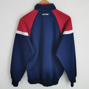 Puede incluir: Una chaqueta deportiva de manga larga con un dise&ntilde;o de bloques de color en azul marino, rojo y blanco. La chaqueta tiene cuello alto, pu&ntilde;os y dobladillo acanalados. Un peque&ntilde;o parche rectangular con el texto FILA est&aacute; centrado bajo el cuello.
