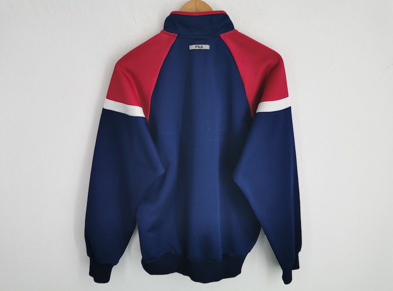 Puede incluir: Una chaqueta deportiva de manga larga con un dise&ntilde;o de bloques de color en azul marino, rojo y blanco. La chaqueta tiene cuello alto, pu&ntilde;os y dobladillo acanalados. Un peque&ntilde;o parche rectangular con el texto FILA est&aacute; centrado bajo el cuello.
