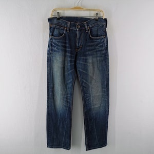 Może przedstawiać: Ciemnoniebieskie dżinsy z denimu wiszące na drewnianym wieszaku. Dżinsy mają klasyczny pięciokieszeniowy design i wyblakły, zużyty wygląd. Denim jest średniej gramatury, a dżinsy wydają się być noszone.