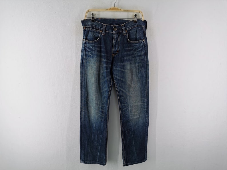 Może przedstawiać: Ciemnoniebieskie dżinsy z denimu wiszące na drewnianym wieszaku. Dżinsy mają klasyczny pięciokieszeniowy design i wyblakły, zużyty wygląd. Denim jest średniej gramatury, a dżinsy wydają się być noszone.