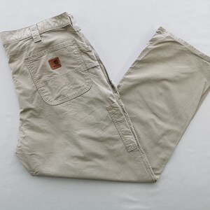 May include: Khaki-colored work pants with multiple pockets and a brown leather patch. The pants are made of a durable material and are designed for work or casual wear. The Carhartt logo is visible on the patch.