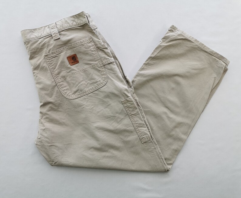May include: Khaki-colored work pants with multiple pockets and a brown leather patch. The pants are made of a durable material and are designed for work or casual wear. The Carhartt logo is visible on the patch.