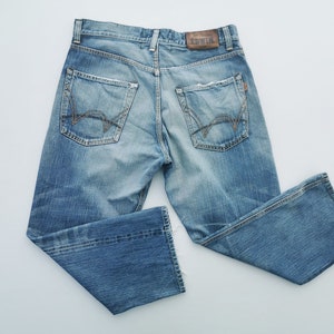 Può includere: Jeans in denim blu chiaro con lavaggio sbiadito. I jeans presentano il classico stile a cinque tasche, chiusura con bottone e cerniera, e una toppa in pelle con il marchio "EDWIN" sul retro. Le tasche posteriori hanno un design ricamato unico.