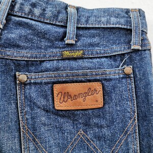 Peut inclure: Gros plan du dos d'un jean Wrangler en denim bleu. Le jean pr&eacute;sente une &eacute;tiquette en cuir marron avec le logo Wrangler sur la poche arri&egrave;re. Les coutures sont de couleur beige contrastante. Le jean a un look classique et vintage.