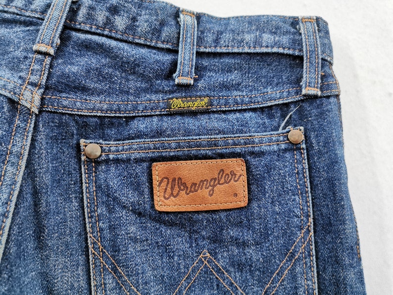 Peut inclure: Gros plan du dos d'un jean Wrangler en denim bleu. Le jean pr&eacute;sente une &eacute;tiquette en cuir marron avec le logo Wrangler sur la poche arri&egrave;re. Les coutures sont de couleur beige contrastante. Le jean a un look classique et vintage.