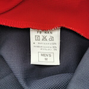 Op de afbeelding: Close-up van een kledingstuk met een rode kraag en een donkerblauwe body. Een wit label toont tekst, waaronder "MEN'S M" en wasvoorschriften. De stof samenstelling is vermeld als polyester en polyurethaan.