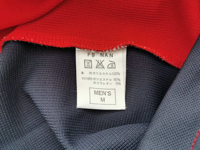 Op de afbeelding: Close-up van een kledingstuk met een rode kraag en een donkerblauwe body. Een wit label toont tekst, waaronder "MEN'S M" en wasvoorschriften. De stof samenstelling is vermeld als polyester en polyurethaan.