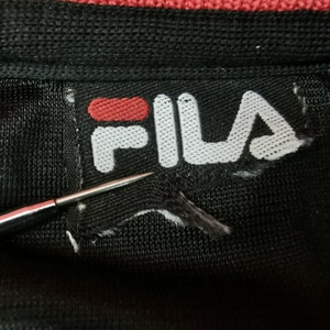 May include: Close-up of a black fabric with a torn white label. The label features the red and white "FILA" logo. A silver needle points towards the label, highlighting the damage. The fabric appears to be part of a garment.