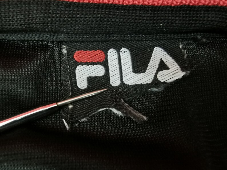 May include: Close-up of a black fabric with a torn white label. The label features the red and white "FILA" logo. A silver needle points towards the label, highlighting the damage. The fabric appears to be part of a garment.