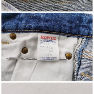 May include: Close-up of the waistband of blue denim jeans. The jeans feature multiple tags with text, including "EDWIN US CLASSIC" and care instructions. The jeans have brass-colored rivets and a classic design.