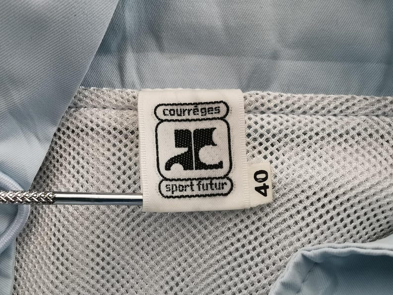May include: Close-up of a white tag with the "courr&egrave;ges sport futur" logo and the number 40. The tag is attached to a light blue garment with a mesh lining. A silver metal object is in the image.