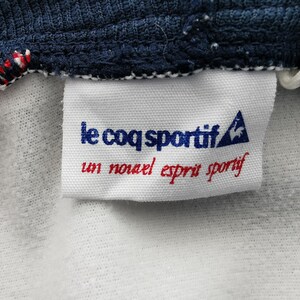 K&ouml;nnte beinhalten: Nahaufnahme eines wei&szlig;en Etiketts mit dem Text "le coq sportif" in Blau und "un nouvel esprit sportif" in Rot. Das Etikett ist an einem marineblauen Stoff mit roten und wei&szlig;en N&auml;hten befestigt.