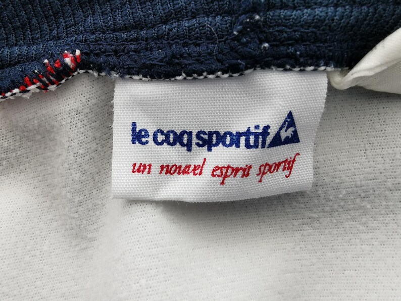 K&ouml;nnte beinhalten: Nahaufnahme eines wei&szlig;en Etiketts mit dem Text "le coq sportif" in Blau und "un nouvel esprit sportif" in Rot. Das Etikett ist an einem marineblauen Stoff mit roten und wei&szlig;en N&auml;hten befestigt.