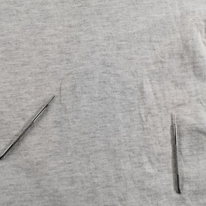 May include: Close-up of a light gray fabric with two silver metal tools. The tools have a pointed end and a textured handle. The fabric has some minor imperfections.