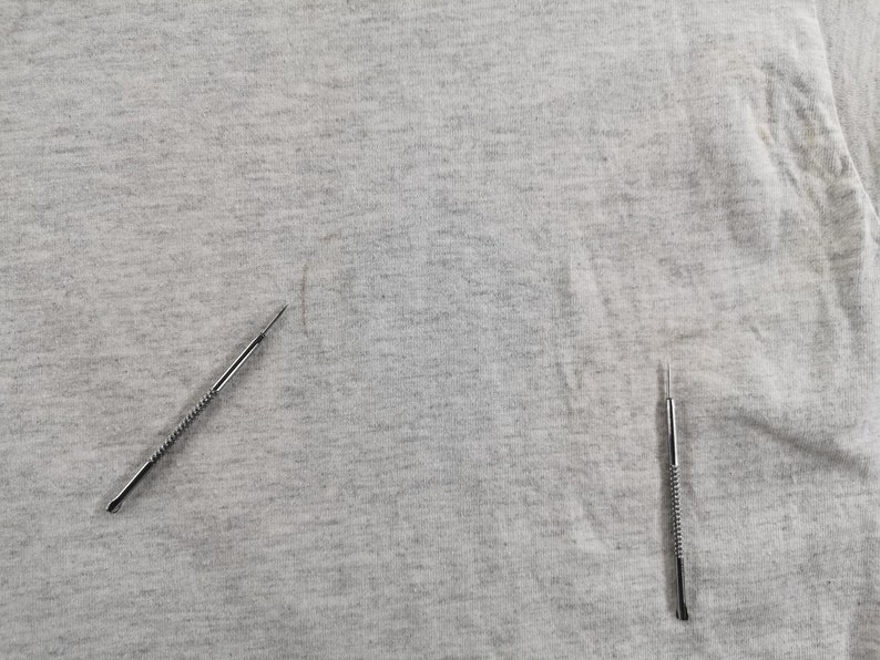 May include: Close-up of a light gray fabric with two silver metal tools. The tools have a pointed end and a textured handle. The fabric has some minor imperfections.