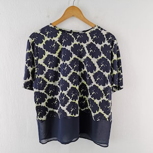 May include: A navy blue top with a floral pattern in white and chartreuse. The bottom panel is a solid navy blue sheer fabric. The top is hanging on a wooden hanger against a white wall.