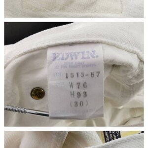 May include: Close-up of white denim fabric with multiple tags. The top tag reads "EDWIN ONLY GENUINE" in blue and black. Other tags show details like "LOT 1513-57" and "W76". The fabric is textured.