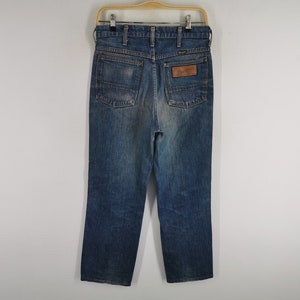 K&ouml;nnte beinhalten: Blaue Jeans aus Denim mit klassischem geradem Bein. Die Jeans haben eine verwaschene Optik, einen braunen Lederaufn&auml;her auf der R&uuml;ckseite und das Wrangler-Logo. Die Jeans h&auml;ngen an einem Holzb&uuml;gel.