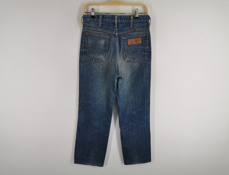 K&ouml;nnte beinhalten: Blaue Jeans aus Denim mit klassischem geradem Bein. Die Jeans haben eine verwaschene Optik, einen braunen Lederaufn&auml;her auf der R&uuml;ckseite und das Wrangler-Logo. Die Jeans h&auml;ngen an einem Holzb&uuml;gel.