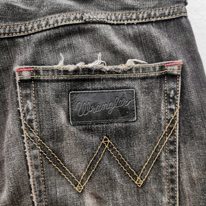 May include: Close-up of the back pocket of black denim jeans. The Wrangler logo is on a black leather patch. The pocket has a signature W-shaped stitching in a contrasting yellow thread. The jeans have a distressed look.