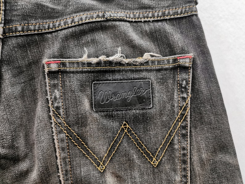 May include: Close-up of the back pocket of black denim jeans. The Wrangler logo is on a black leather patch. The pocket has a signature W-shaped stitching in a contrasting yellow thread. The jeans have a distressed look.