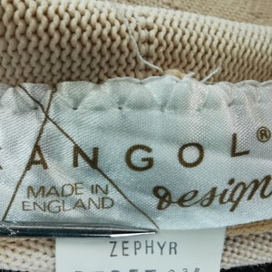 Puede incluir: Primer plano de una tela de punto beige con una etiqueta que dice "KANGOL design MADE IN ENGLAND". La etiqueta tambi&eacute;n tiene la palabra "ZEPHYR" impresa. La tela parece ser parte de un sombrero u otro tocado.