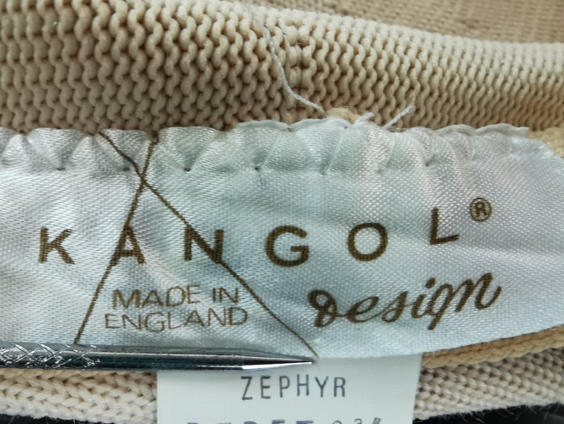 Puede incluir: Primer plano de una tela de punto beige con una etiqueta que dice "KANGOL design MADE IN ENGLAND". La etiqueta tambi&eacute;n tiene la palabra "ZEPHYR" impresa. La tela parece ser parte de un sombrero u otro tocado.