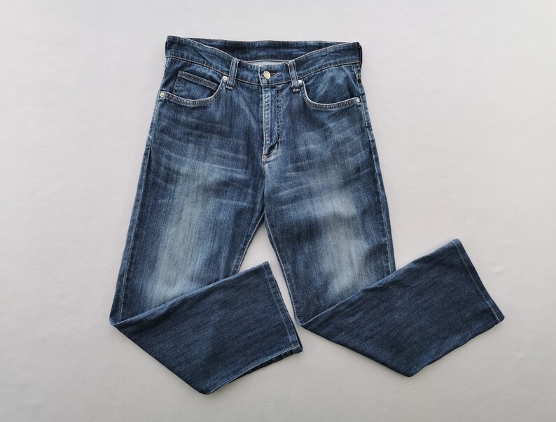 May include: Dark blue denim jeans with a classic five-pocket design. The jeans feature a button and zip fastening, belt loops, and a faded wash. The jeans are displayed flat on a grey surface.