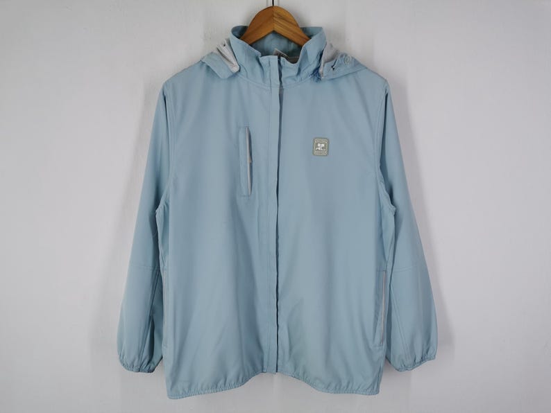 May include: A light blue hooded jacket with a full zipper closure. The jacket has a small square logo on the chest and a zippered pocket on the left side. The jacket is hanging on a wooden hanger.