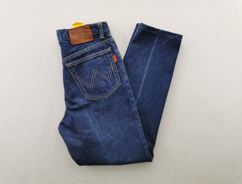 May include: Dark blue denim jeans with a classic five-pocket design. Features a brown leather patch with embossed text and a distinctive W-shaped stitching on the back pockets. The jeans have a straight leg cut and a small red tag.