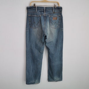 May include: Light blue denim jeans hanging on a wooden hanger. The jeans have a classic five-pocket design, a brown leather patch with the "Wrangler" logo, and a faded wash. The jeans are a straight-leg style.