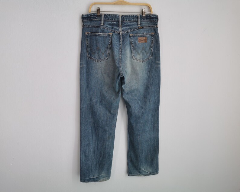May include: Light blue denim jeans hanging on a wooden hanger. The jeans have a classic five-pocket design, a brown leather patch with the "Wrangler" logo, and a faded wash. The jeans are a straight-leg style.
