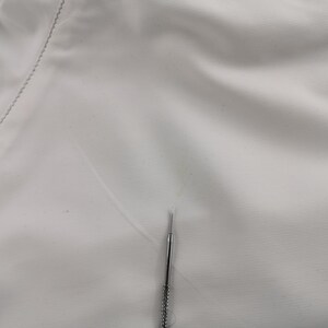 Puede incluir: Primer plano de una tela blanca con una costura visible en el lado izquierdo. Una aguja plateada est&aacute; posicionada verticalmente en el centro, apuntando hacia arriba. La tela tiene una textura sutil.