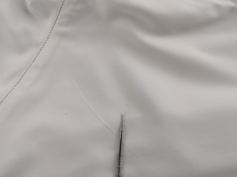 Puede incluir: Primer plano de una tela blanca con una costura visible en el lado izquierdo. Una aguja plateada est&aacute; posicionada verticalmente en el centro, apuntando hacia arriba. La tela tiene una textura sutil.