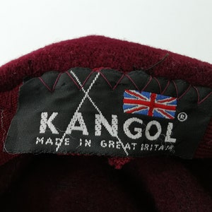 May include: Close-up of a burgundy Kangol hat label. The black label features the brand name, the Union Jack flag, and the text "MADE IN GREAT BRITAIN". The hat's material is visible.