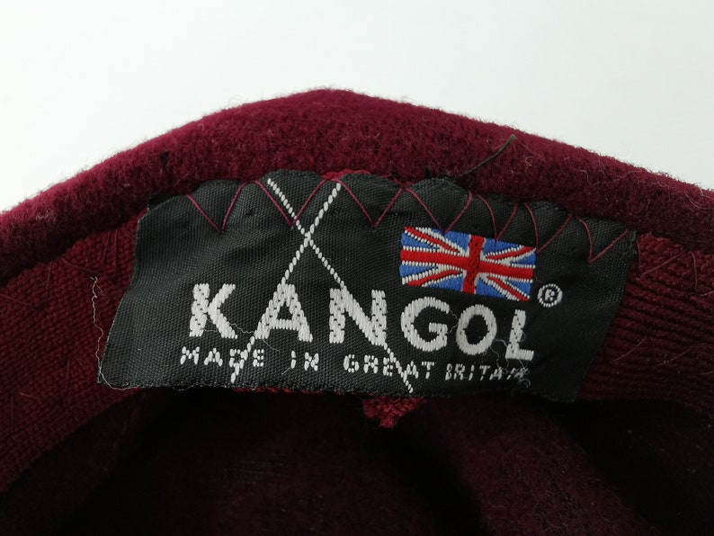 May include: Close-up of a burgundy Kangol hat label. The black label features the brand name, the Union Jack flag, and the text "MADE IN GREAT BRITAIN". The hat's material is visible.