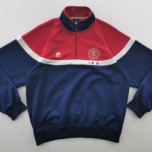 Puede incluir: Una chaqueta deportiva roja y azul marino con cremallera corta, cuello alto, mangas largas con pu&ntilde;os acanalados y dobladillo acanalado. El pecho muestra un logotipo Fila blanco, un emblema circular y el texto 工藤 楓 sobre una franja blanca.