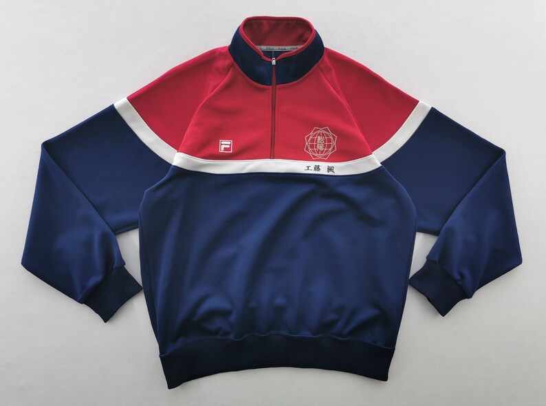 Puede incluir: Una chaqueta deportiva roja y azul marino con cremallera corta, cuello alto, mangas largas con pu&ntilde;os acanalados y dobladillo acanalado. El pecho muestra un logotipo Fila blanco, un emblema circular y el texto 工藤 楓 sobre una franja blanca.