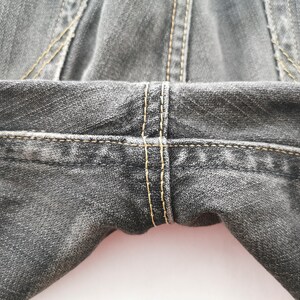 May include: Close-up of the waist and upper thigh area of dark gray denim jeans. The jeans have visible yellow stitching along the seams and around the pockets. The fabric has a textured appearance.